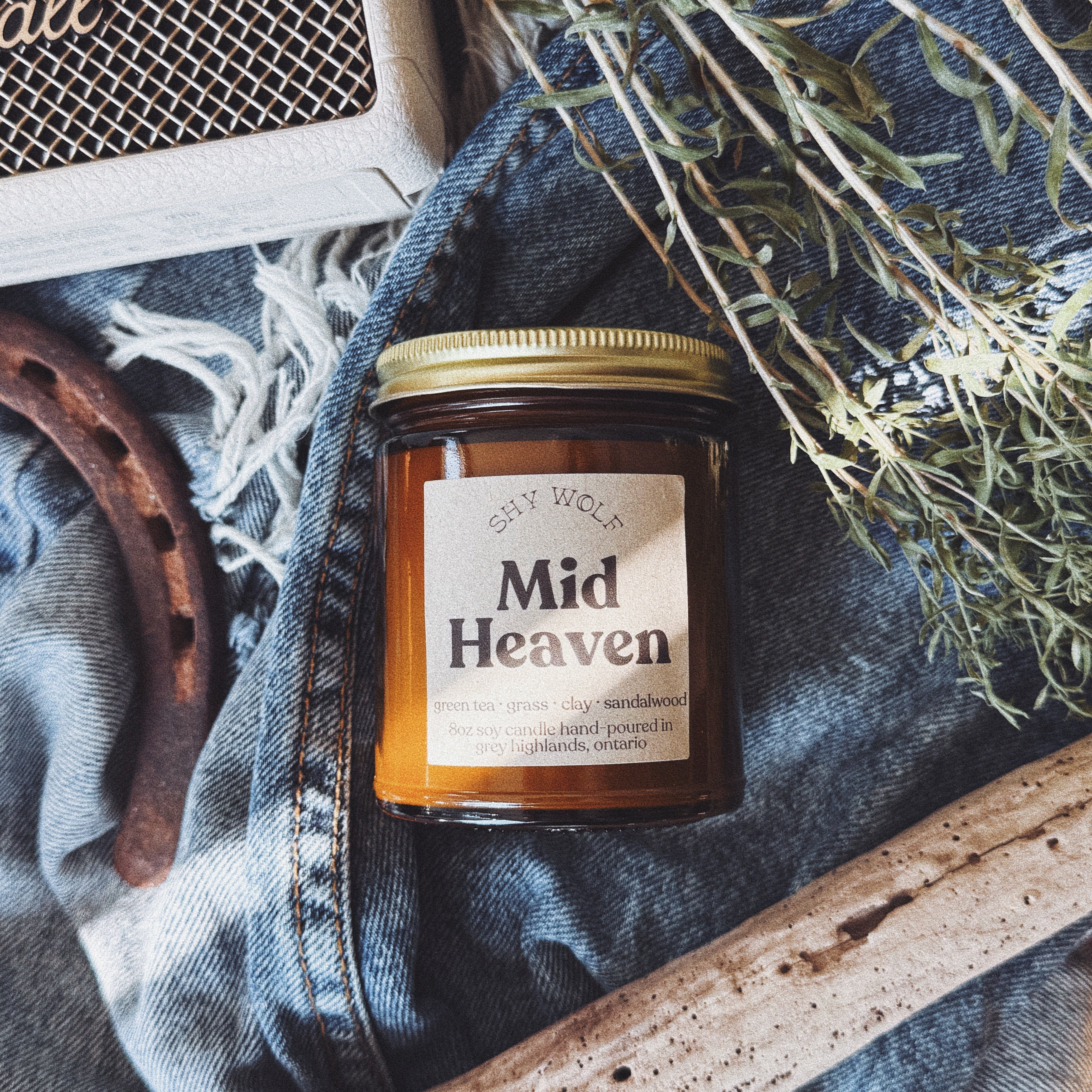 A soy wax candle in a clear glass jar with amber-colored wax and a gold lid, labeled 'Mid Heaven' by Sky Wolf. The jar is styled in a rustic flat-lay arrangement on blue denim with dried herbs, a horseshoe, and a wooden branch.