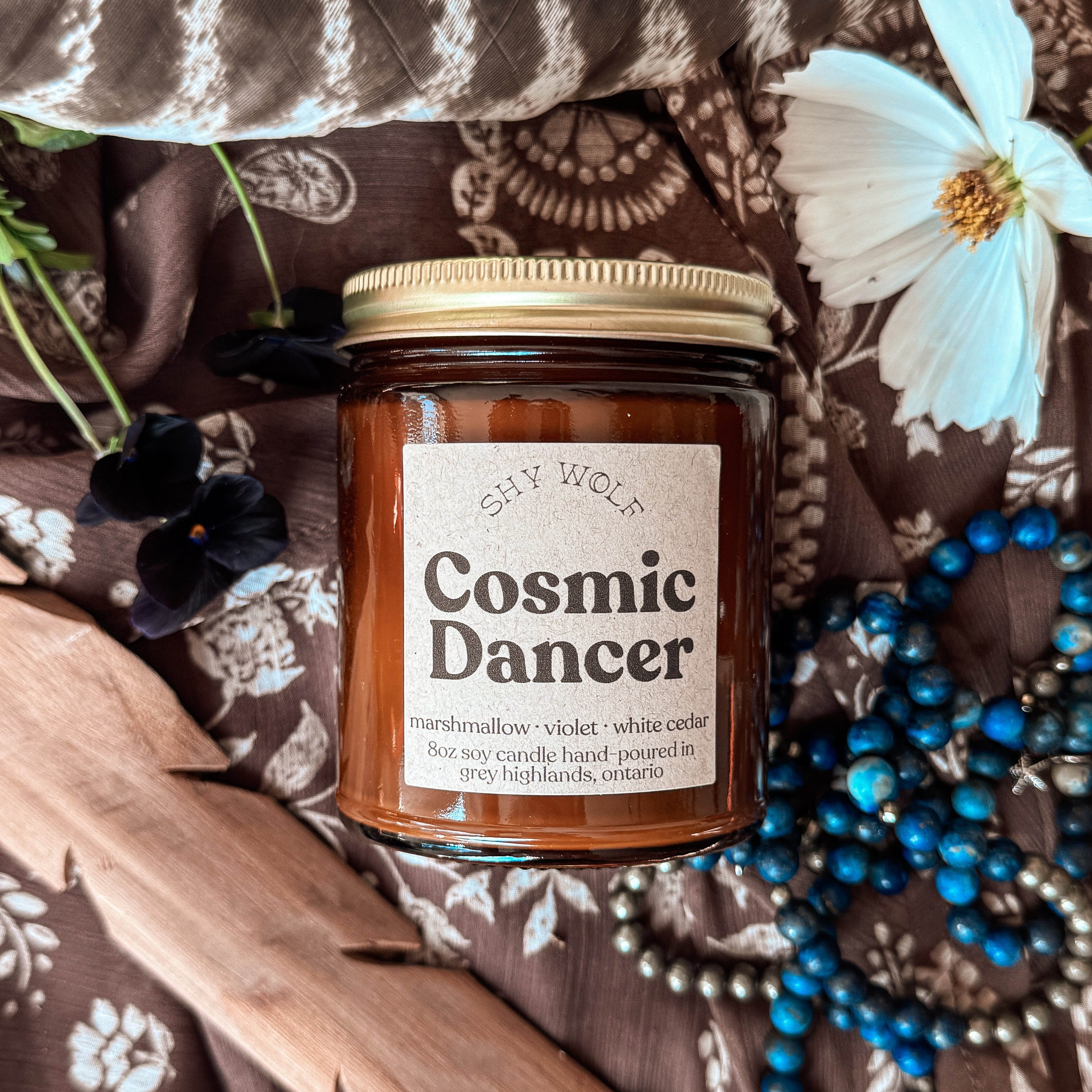 A glass jar candle labeled 'Cosmic Dance' by 'The Witch Cafe' is styled in a flat-lay arrangement on a brown and white patterned fabric with a wooden leaf, blue beads, a white flower, and a feather.