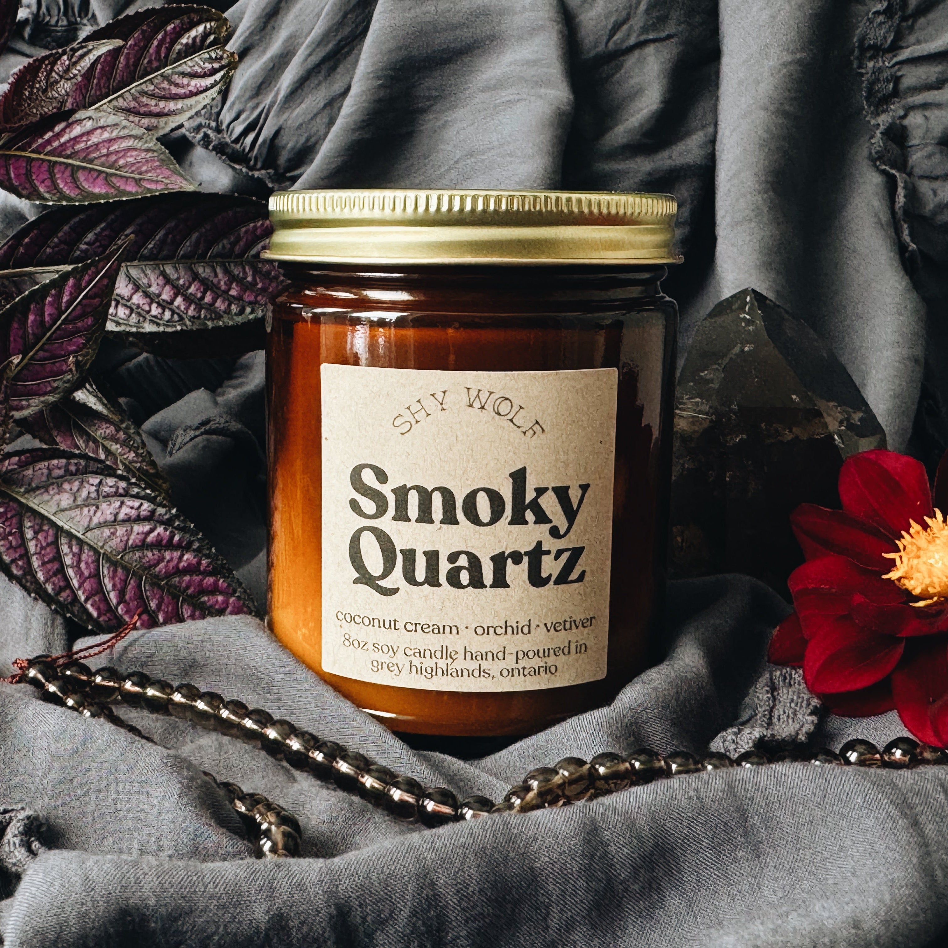 A glass jar candle labeled 'Shy Wolf Smoky Quartz' sits on draped gray fabric, surrounded by dark purple leaves, a red flower, and a crystal cluster.