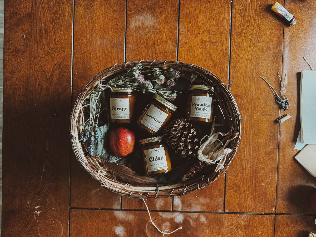 A top-down view of a rustic wicker basket filled with four amber glass jars of spices or seasonings, alongside dried flowers, a red apple, a pinecone, and autumn leaves, arranged on a warm wooden tabletop.